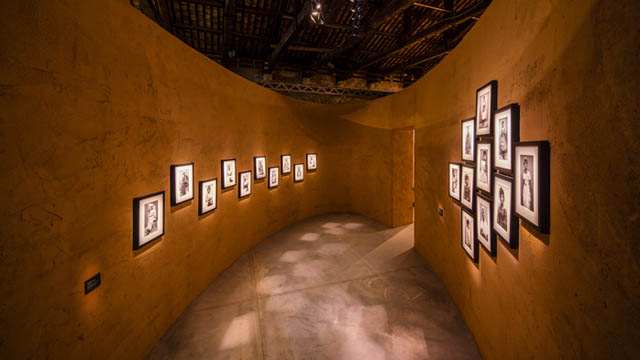 Die Fotographie im Querformat wurde im Ghana-Pavillon auf der Venedig-Biennale 2019 aufgenommen. Der Blick in einen Raum mit gekurvten ockerfarbenen Wänden. Der Raum hat einen ovalen Grundriss. Der Raum hat keine Decke, das offene Dachwerk liegt nicht auf den Wänden auf. An den Wänden hängen gerahmte schwarz-weiß Photographien der Photographin Feclicia Abban.